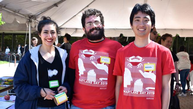 Three Linguistics students, wearing Maryland Day t-shirts, arm in arm and smiling at the camera, doing outreach activities for Maryland Day 2019